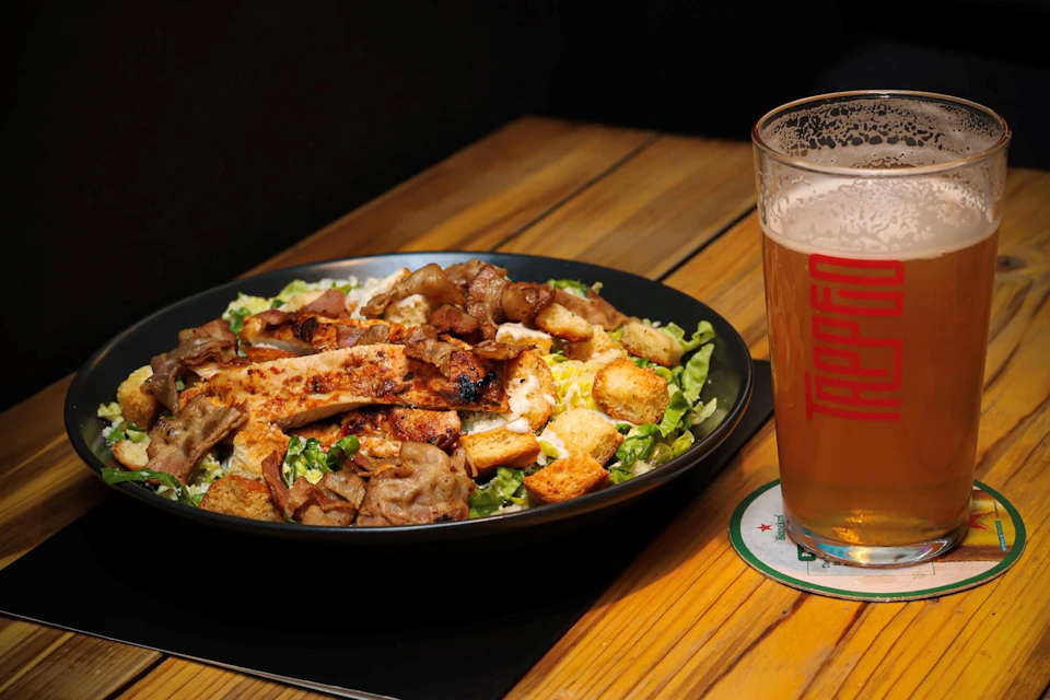 A bowl of salad with grilled chicken and croutons on a wooden table next to a glass of beer.