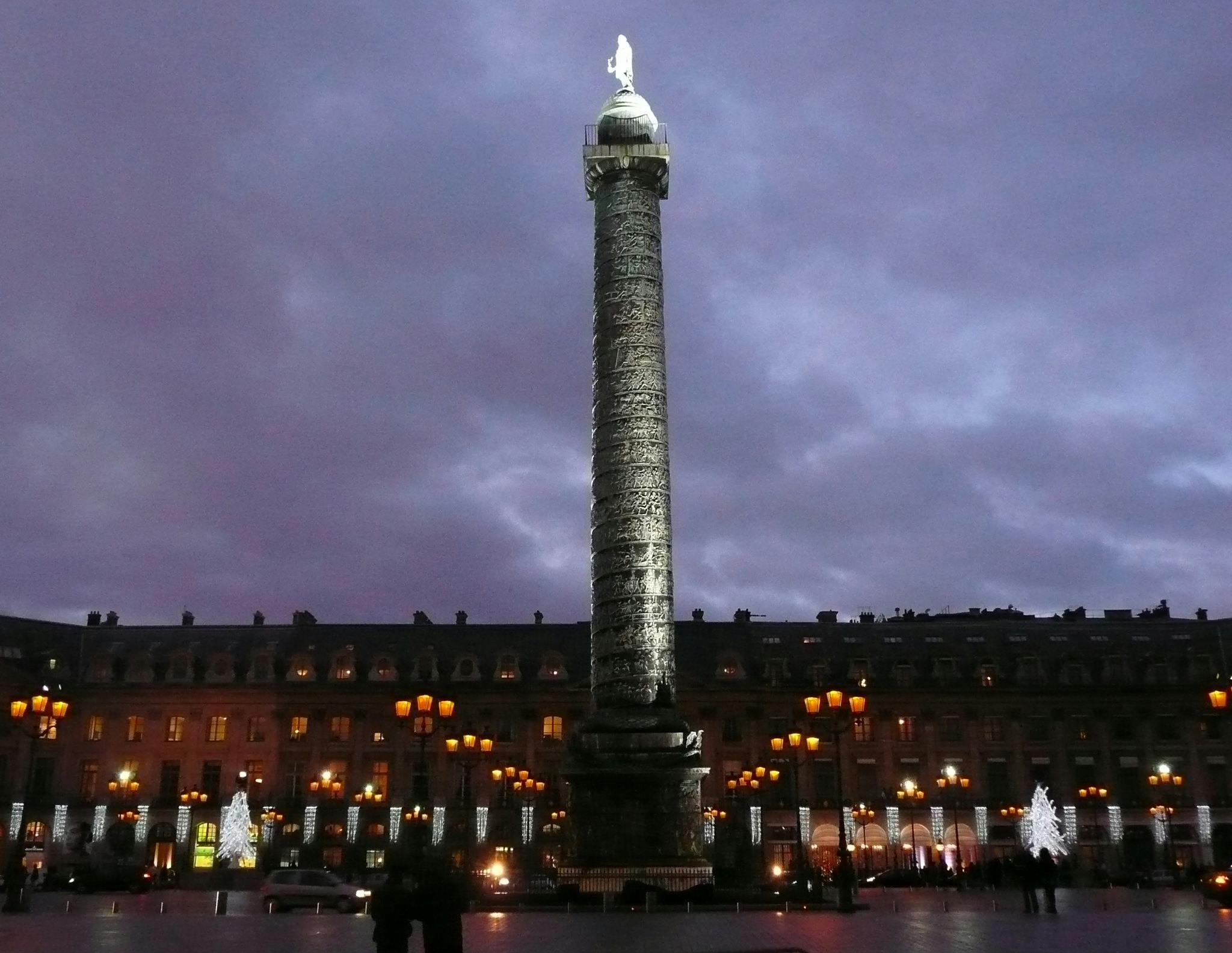 Duża kolumna z posągiem na górze, otoczona oświetlonymi budynkami i latarniami, na tle ciemnego zachmurzonego nieba.