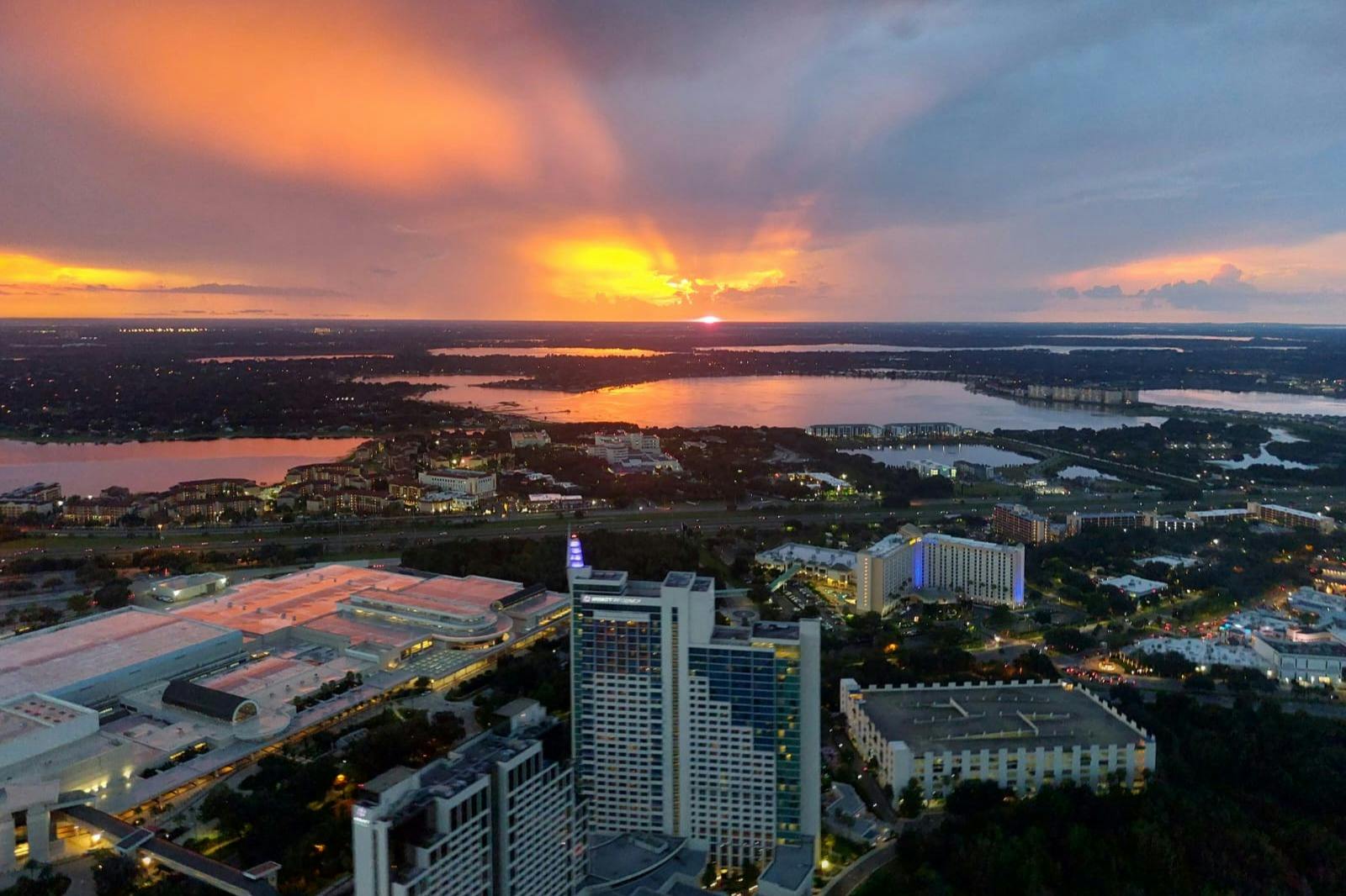 Aerial view of a cityscape at sunset with buildings, a large lake, and a dramatic orange and purple sky.
