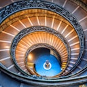 Scala a chiocciola con ringhiera ornata, che scende in modo circolare e conduce a un pavimento di piastrelle blu con una colonna decorativa centrale.