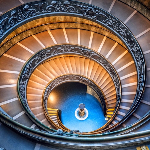 Scala a chiocciola con ringhiera ornata, che scende in modo circolare e conduce a un pavimento di piastrelle blu con una colonna decorativa centrale.