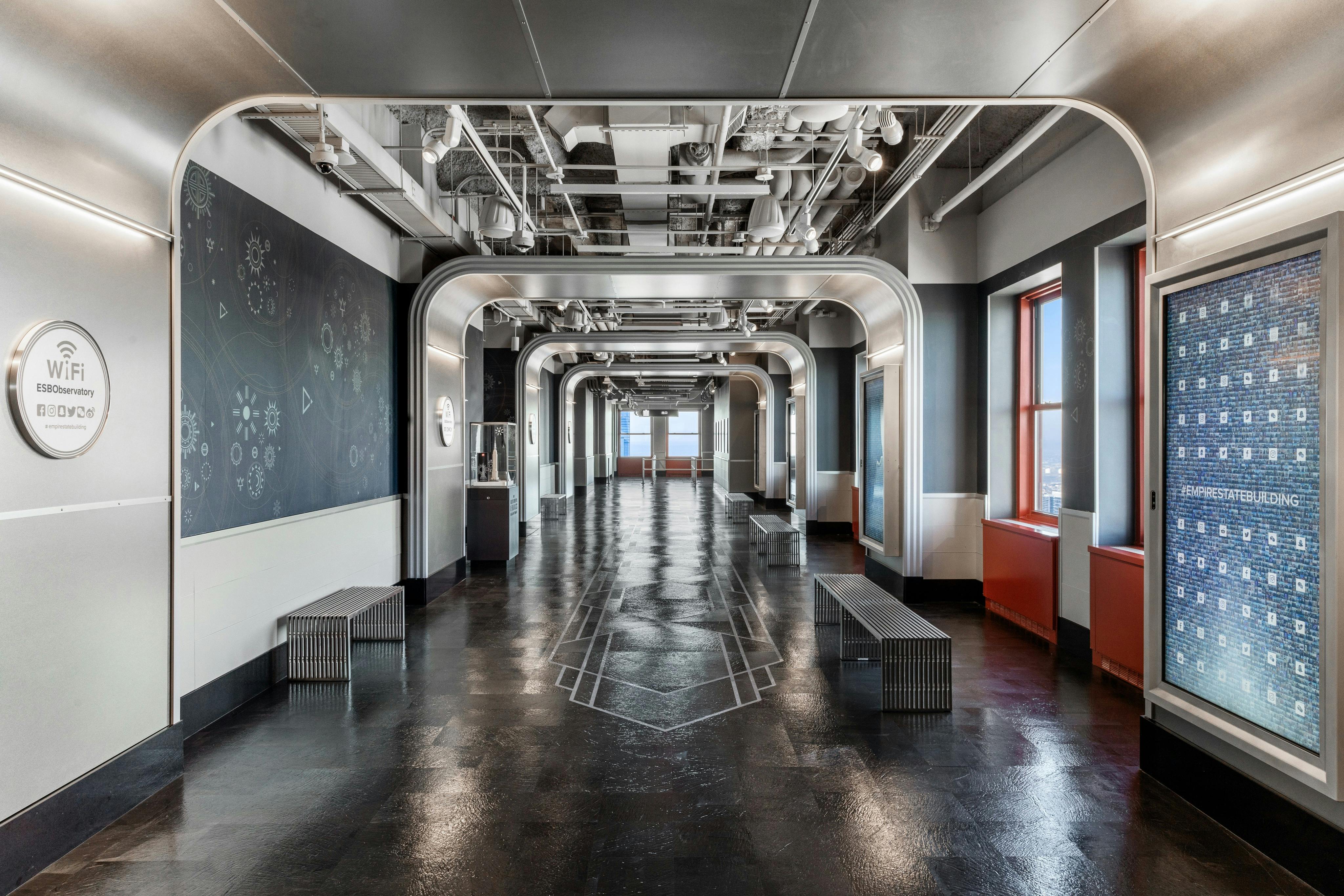 Современная прихожая в индустриальном стиле с арочными металлическими рамами, светоотражающим напольным покрытием, скамейками с каждой стороны и окнами справа.