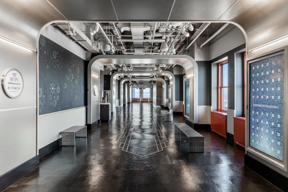 A modern, industrial-style hallway with arched metal frames, reflective flooring, benches on each side, and windows on the right.