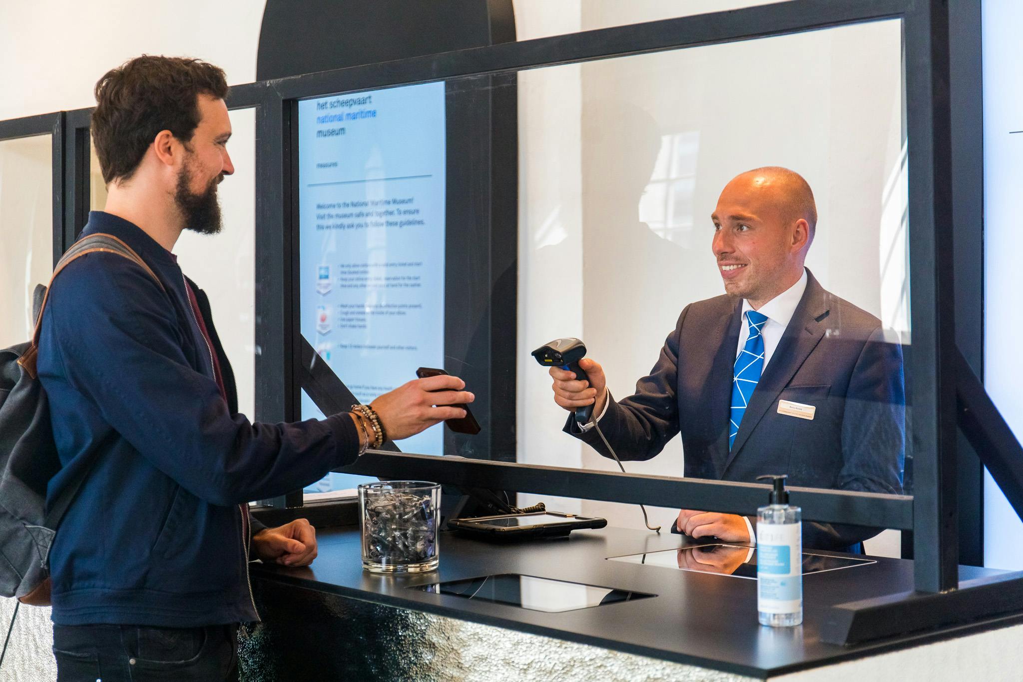 A hotel receptionist in a suit and blue tie uses a scanner behind a glass barrier. A hand from the other side holds a phone.