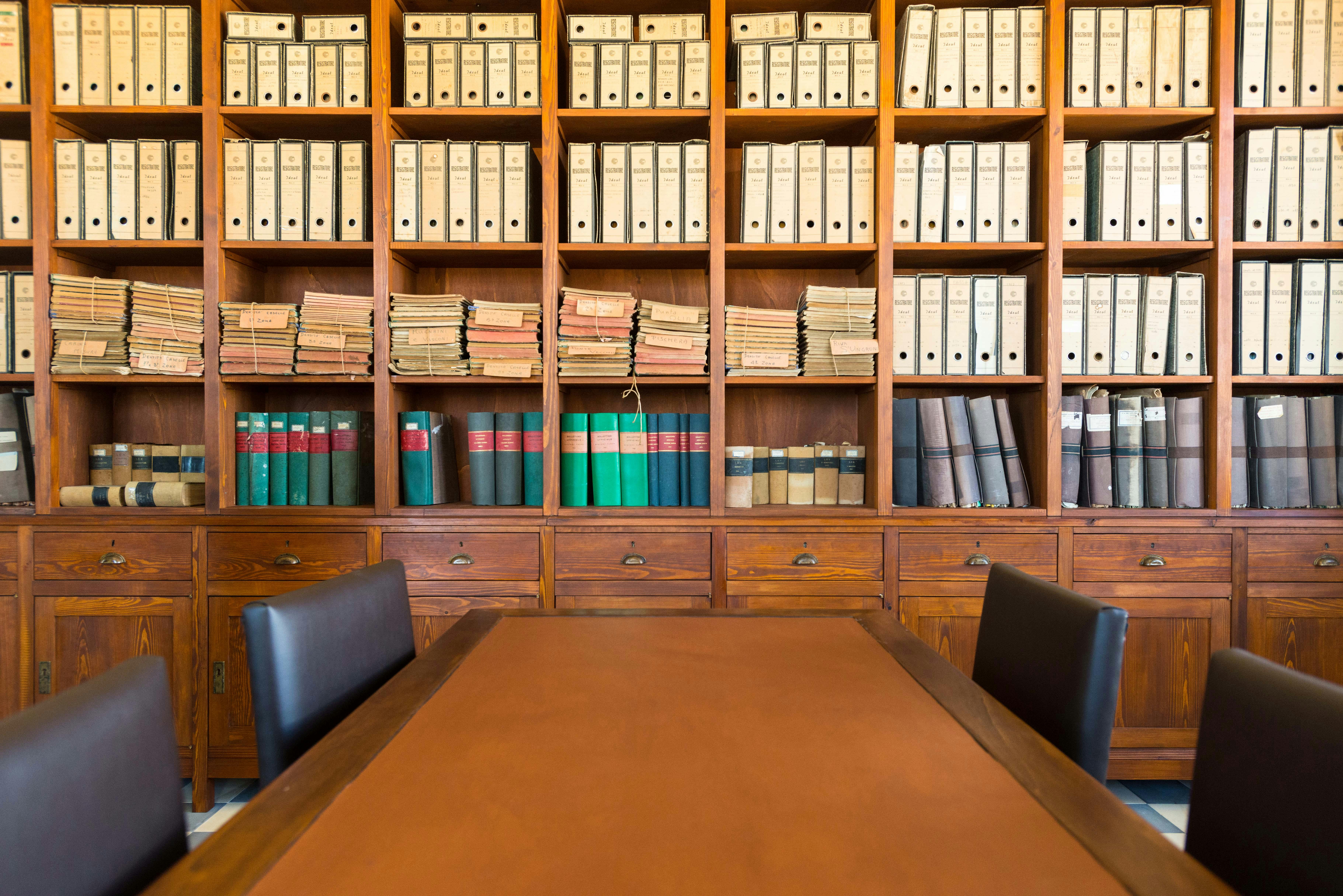Træhylder fyldt med ringbind, bøger og filer bag et konferencebord med to sorte stole.