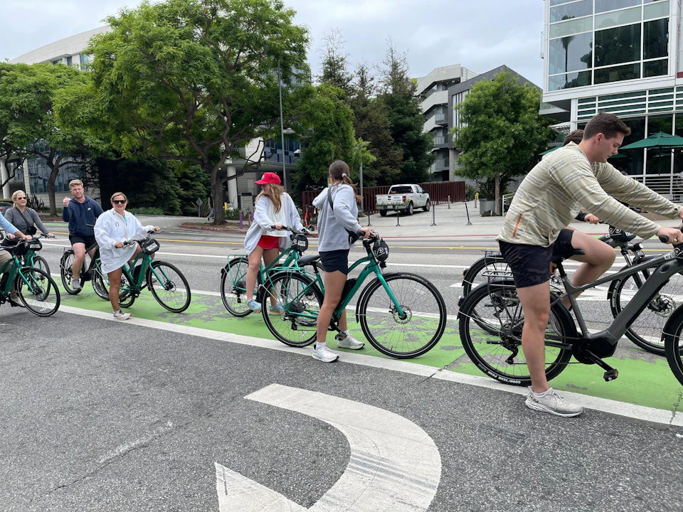 Group of cyclists riding green bikes on a city street with green bike lanes, trees, and buildings in the background.