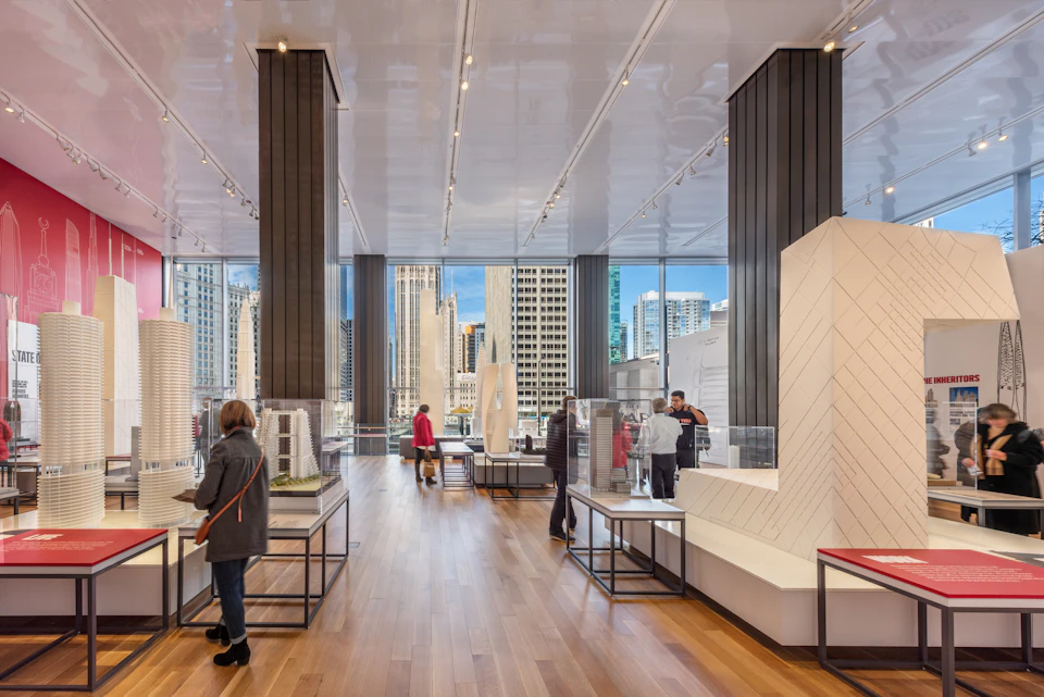 People viewing architectural models in a spacious, modern gallery with large windows overlooking a city skyline.