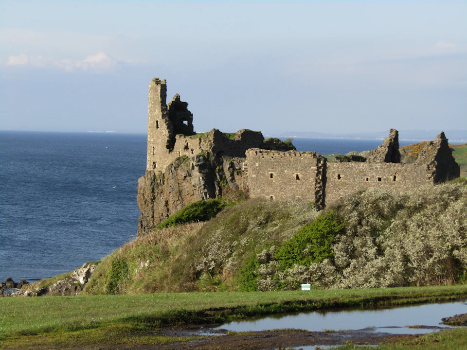 Ruins of a stone castle perched on a grassy cliff overlooking the ocean, with clear skies in the background.
