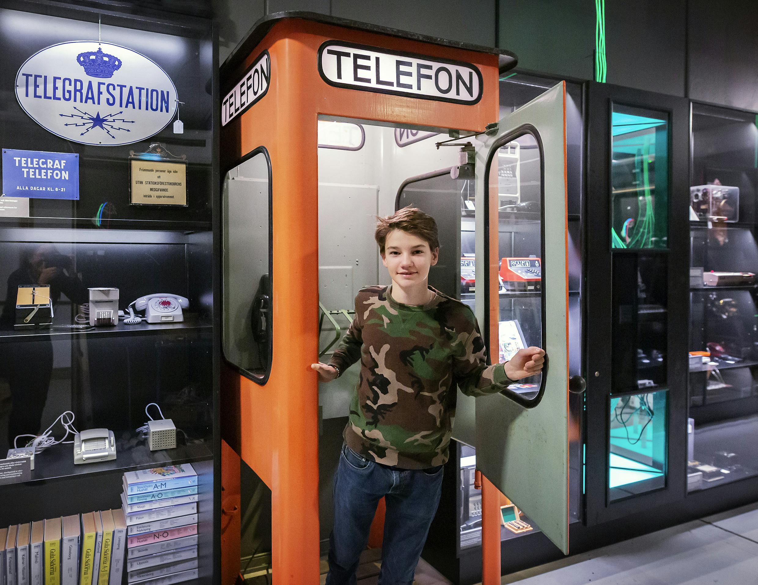 V oranžové telefonní budce s nápisem „TELEFON“ stojí člověk obklopený vitrínami se starožitnou elektronikou.