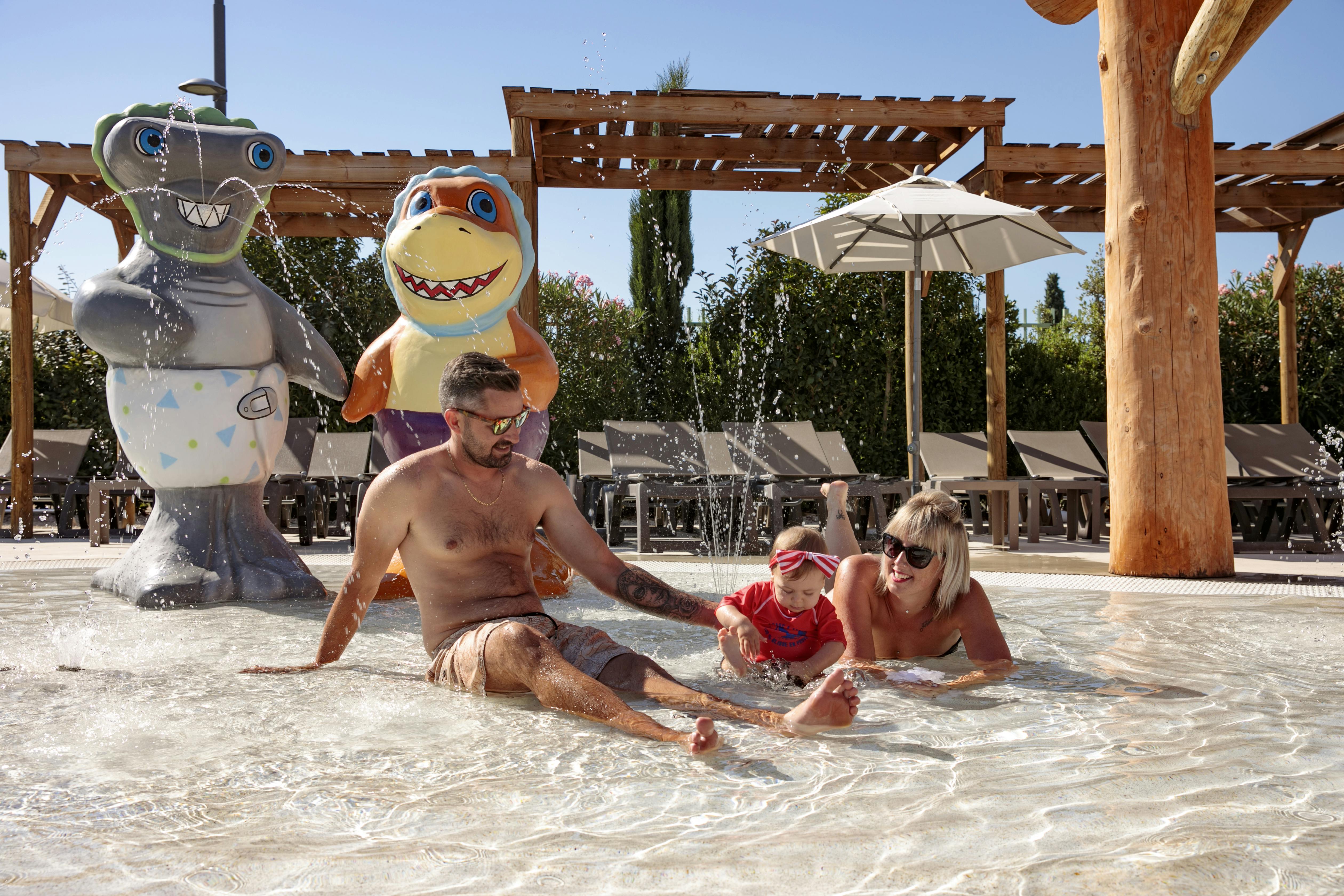 A man, woman, and child play in a shallow pool with water jets, surrounded by cartoon character statues and lounge chairs.