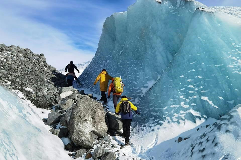 Four hikers in colorful jackets ascend a rocky, snowy path next to a large, translucent blue ice wall under a clear sky.