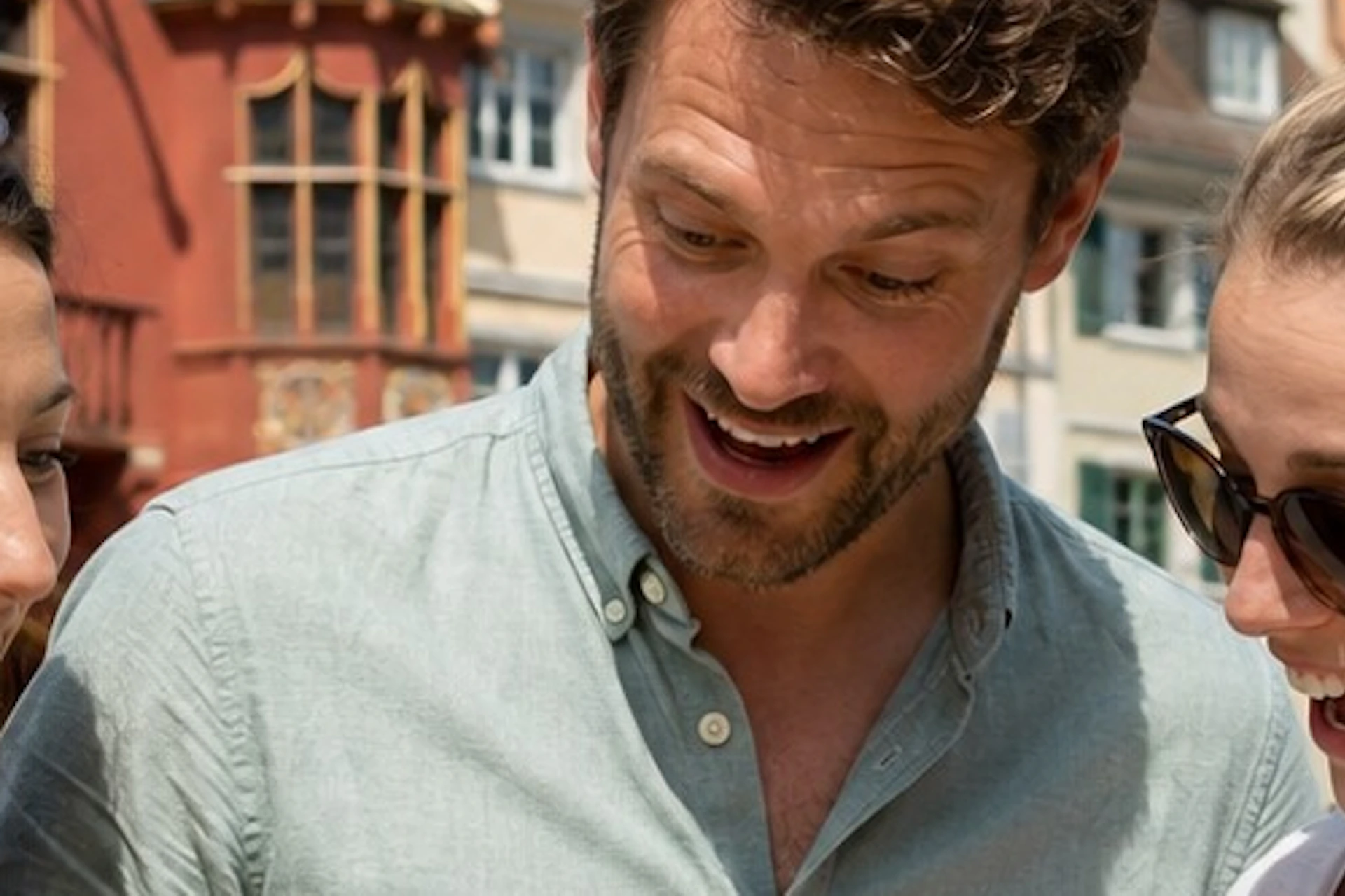 Three people outdoors smiling and looking at a smartphone held by a man, with historic buildings in the background.