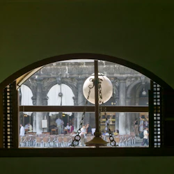 Välvda fönster med utsikt över en innergård med människor, uteserveringsstolar och vita baldakiner på en historisk stenbyggnad.