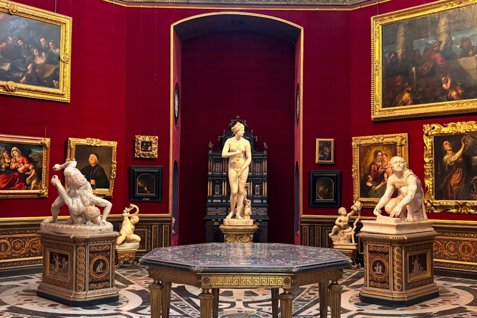 A museum gallery with red walls displaying classical sculptures and framed paintings, centered around a marble statue of Venus.