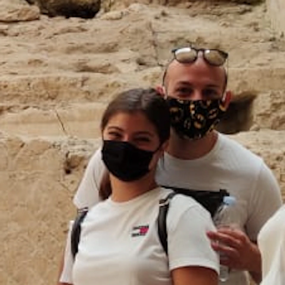 A group of people wearing masks stands on a ramp in front of ancient ruins with painted walls and an archway.