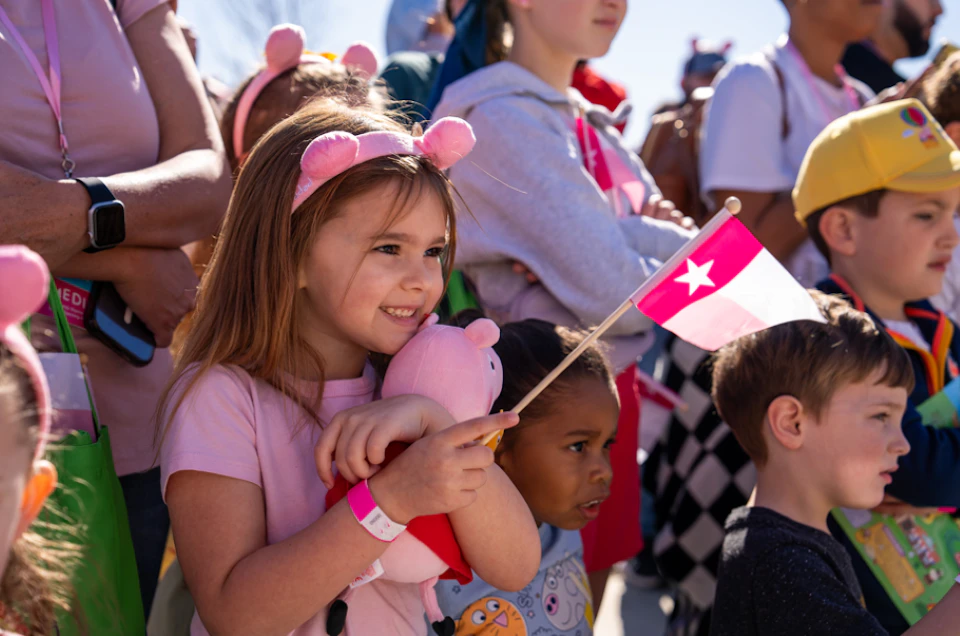 Children standing in a crowd, one girl holding a pink flag and stuffed toy. Bright, sunny day with people in the background.