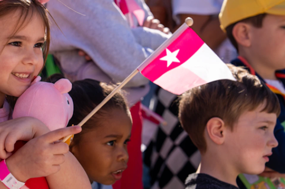 Children standing in a crowd, one girl holding a pink flag and stuffed toy. Bright, sunny day with people in the background.