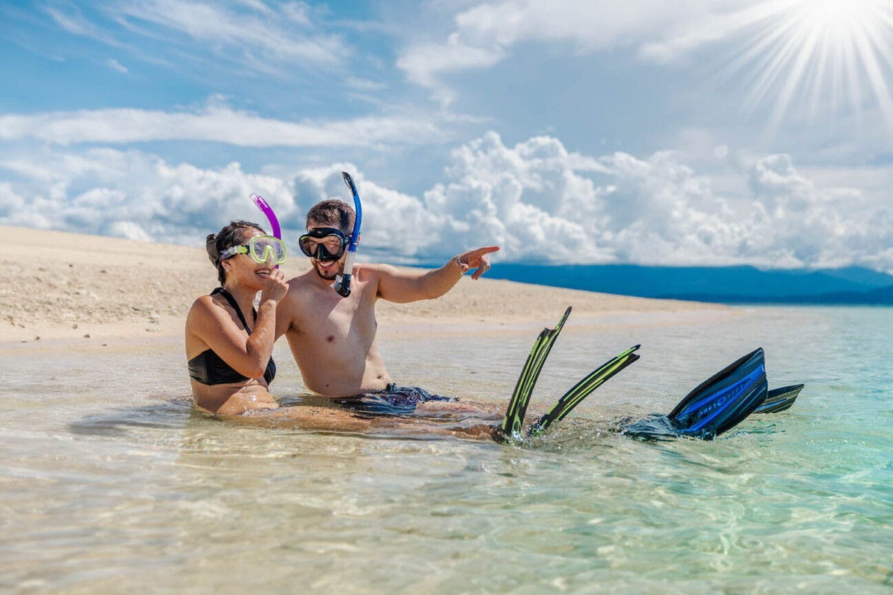Een man en vrouw in snorkeluitrusting zitten in ondiep, helder water op een strand, waarbij de man in de verte wijst.