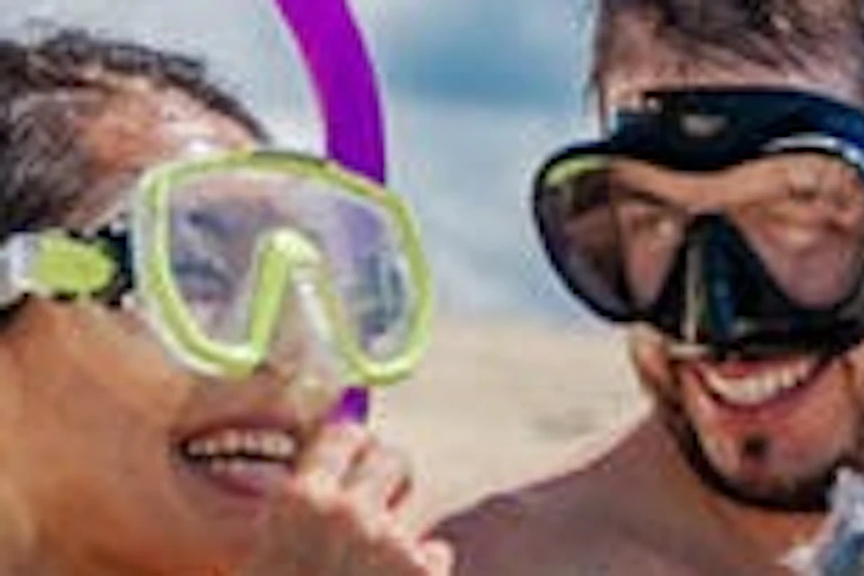 A man and woman in snorkeling gear sit in shallow, clear water on a beach, with the man pointing into the distance.