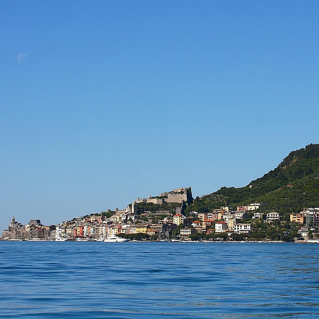Coastal town with colorful buildings, a hill with greenery, and a clear blue sky. Calm water in the foreground.