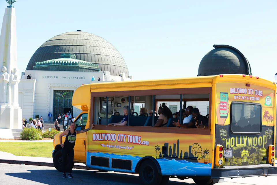 A yellow Hollywood City Tours bus parked near the Griffith Observatory with tourists inside. A guide stands outside the bus.