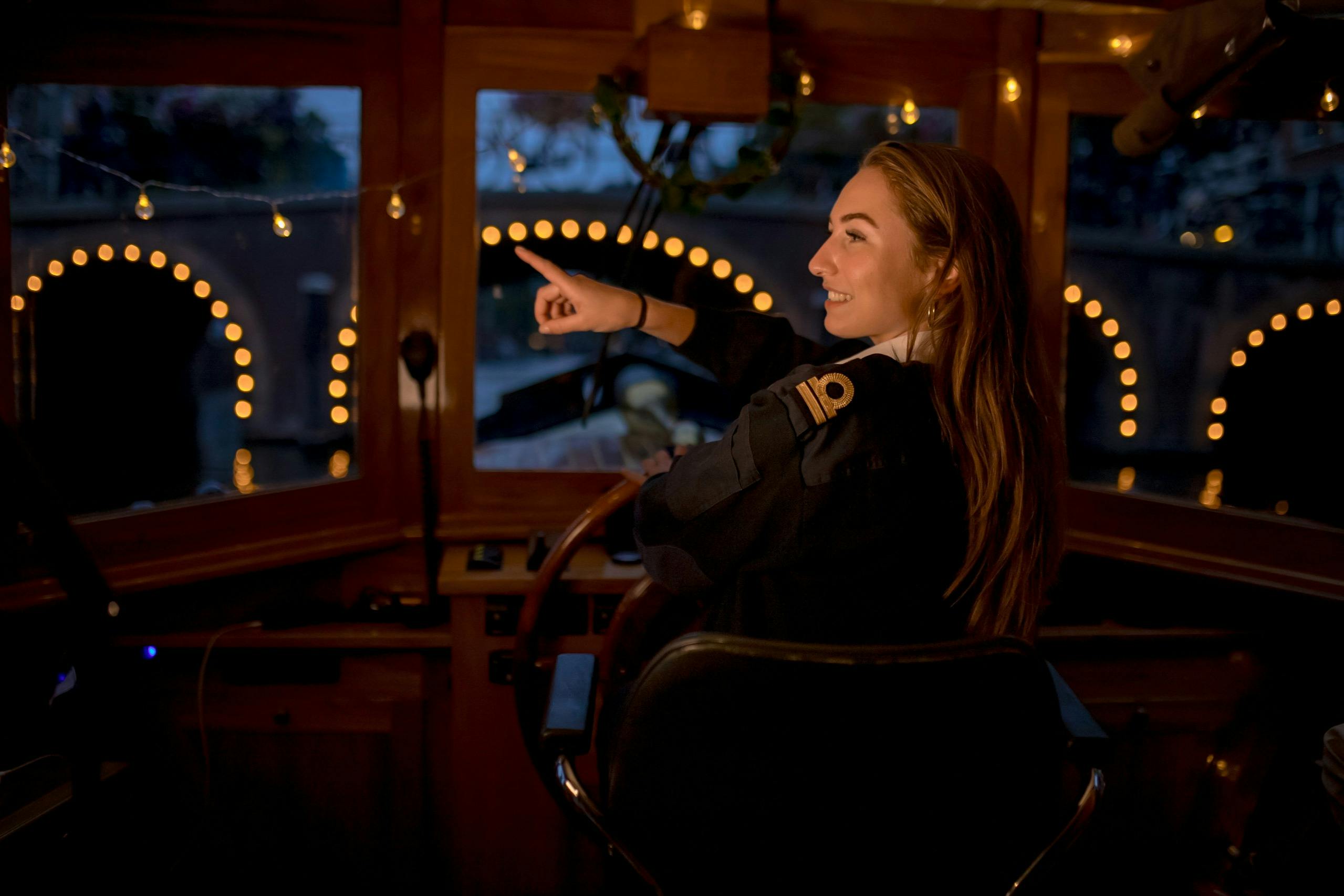 Eine Frau in Uniform steuert ein nächtliches Boot und zeigt nach vorn, wobei durch das Fenster festliche Lichter und Wasserspiegelungen zu sehen sind.