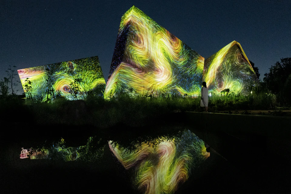 Buildings with swirling light projections resembling Van Gogh's style, reflected in a pool, under a dark sky.