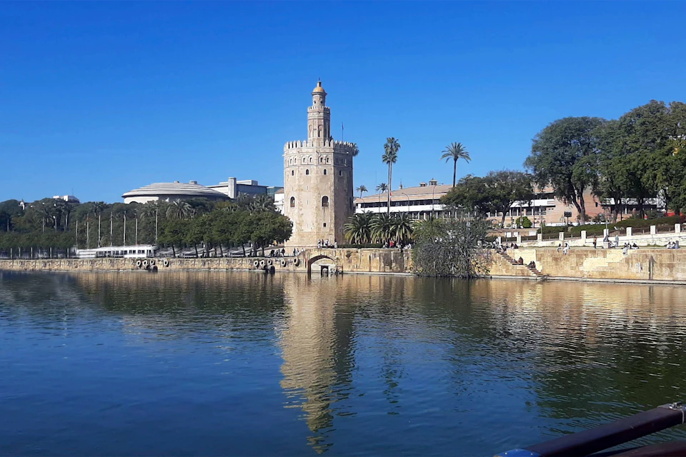 A historic cylindrical tower by a river, surrounded by trees and buildings under a clear blue sky.