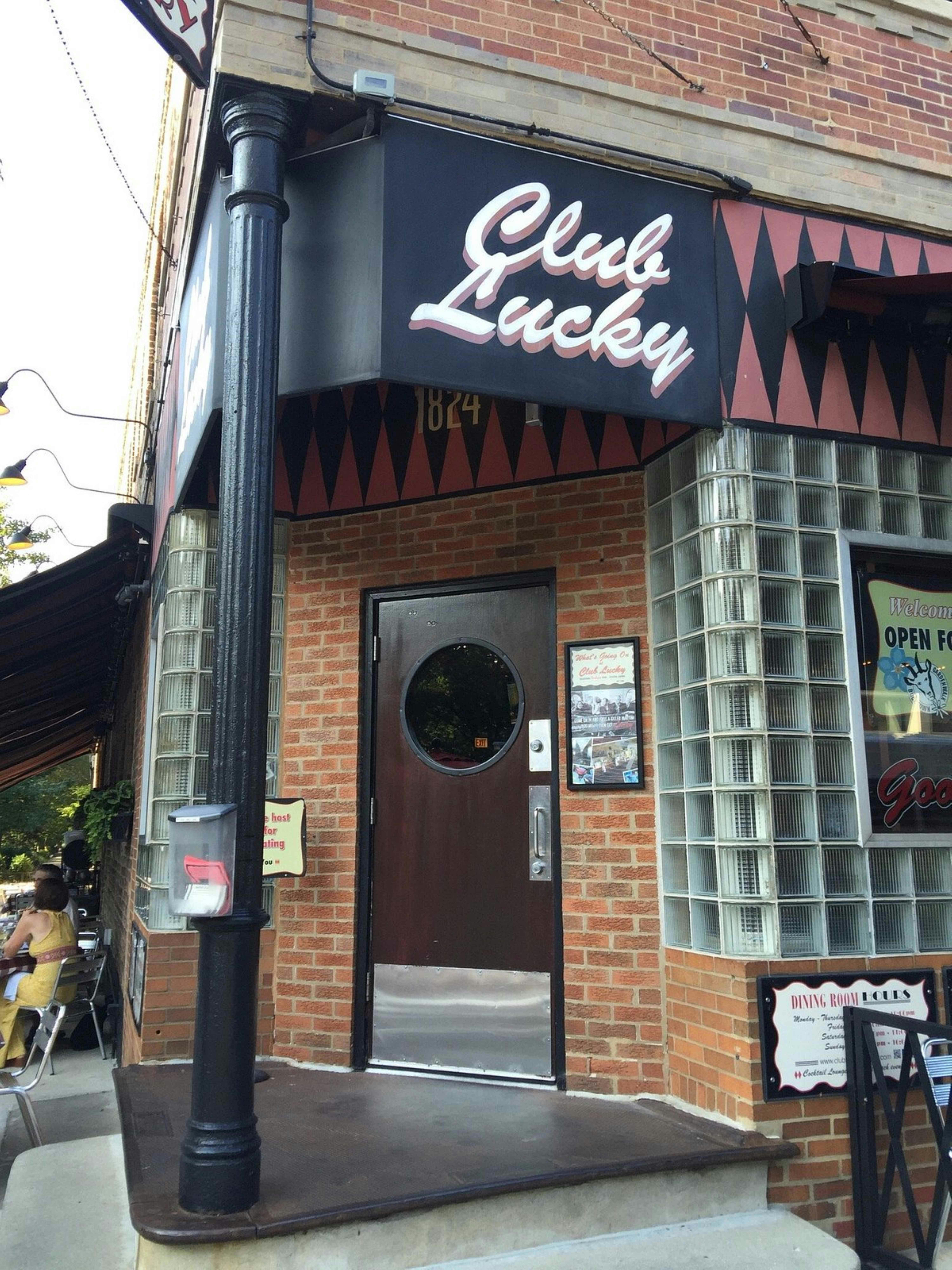 Brick building entrance with a round-windowed door under a "Club Lucky" sign. People are seated at outdoor tables to the left.