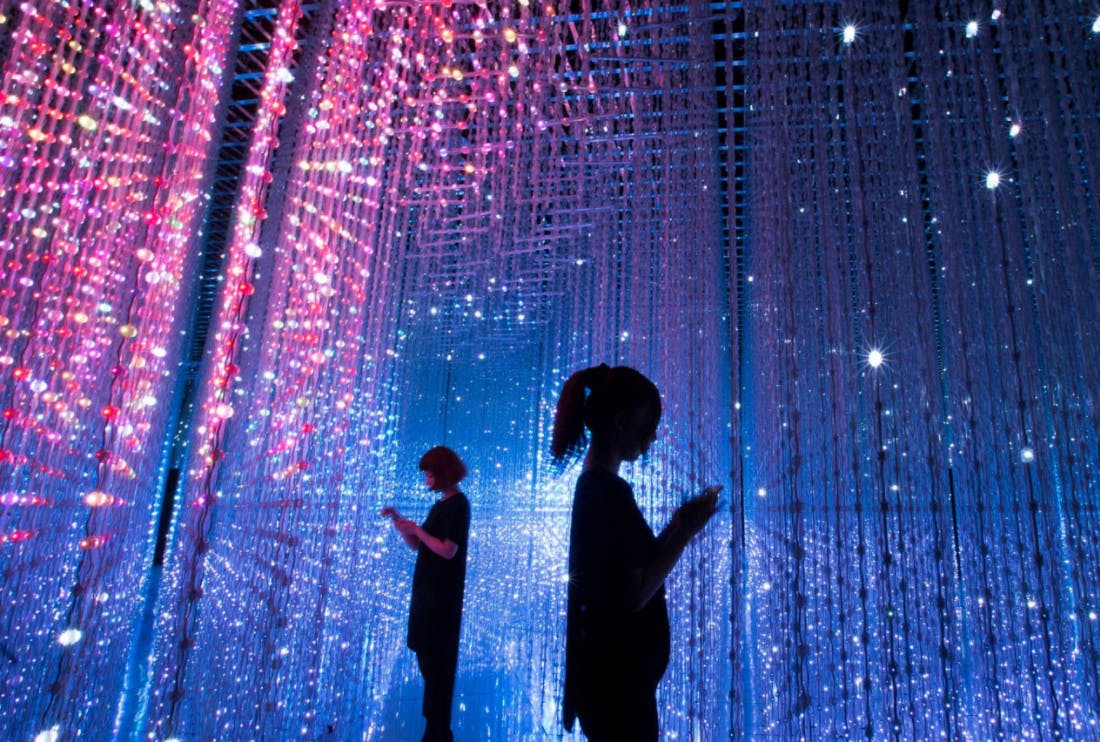 Dues persones s'aixequen enmig de fils penjats de llums multicolors en un espai fosc, immersiu i futurista.
