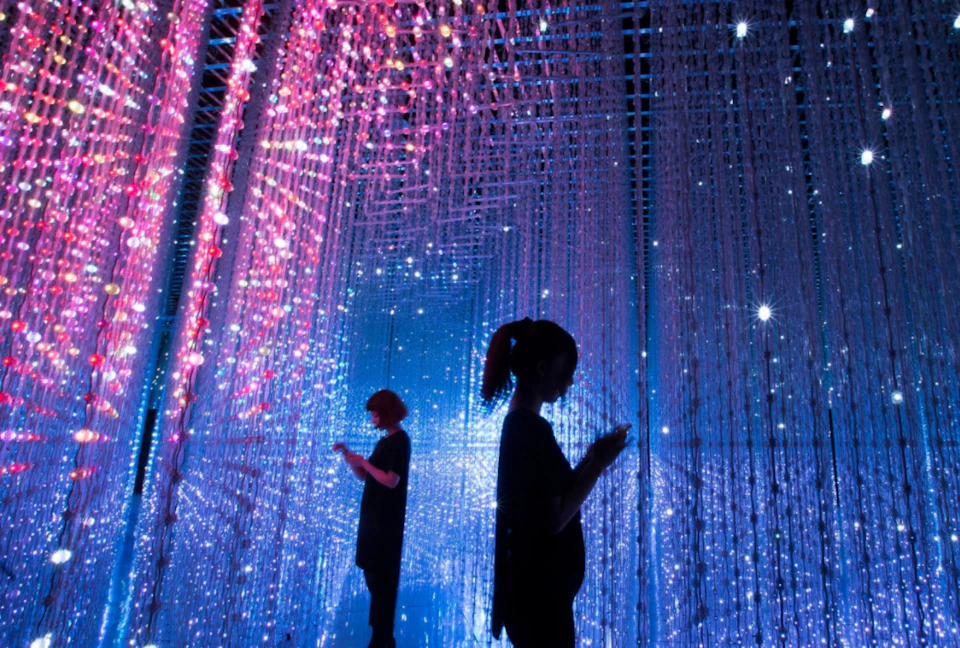 Two people stand amidst hanging strands of multicolored lights in a dark, immersive, and futuristic space.