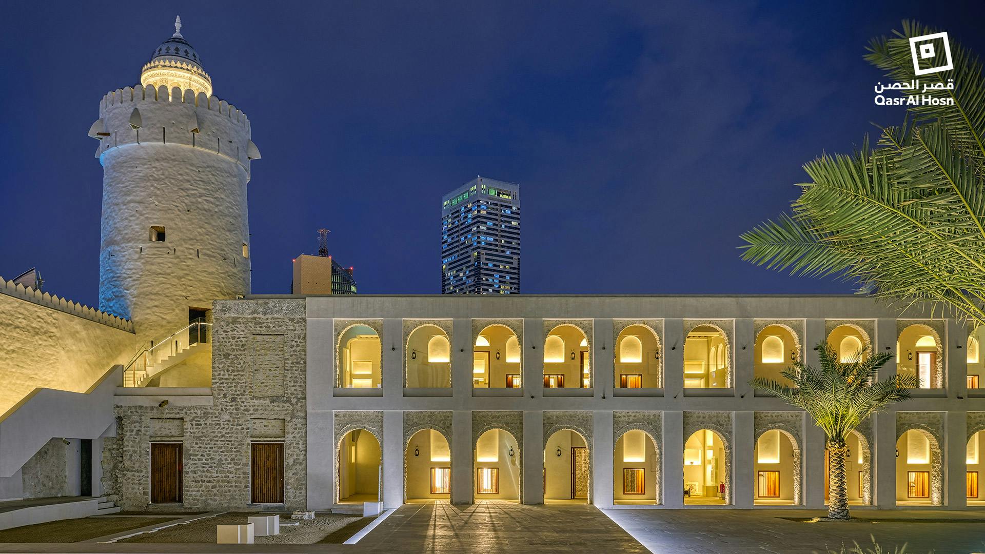 A courtyard with an illuminated two-story building featuring arches at night, with modern high-rises in the background.