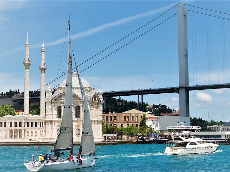 Sailboats on a turquoise sea with a mosque, buildings, and a suspension bridge in the background under a clear blue sky.