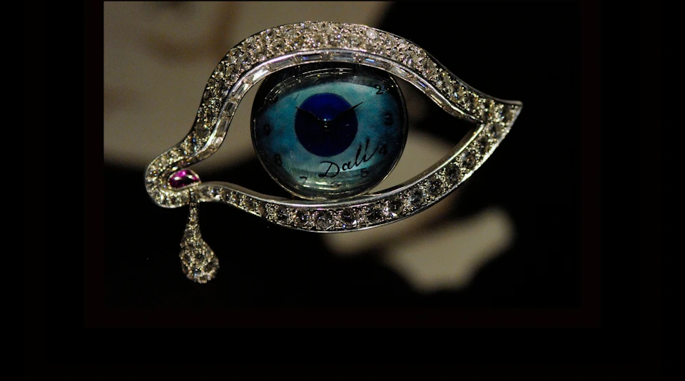 Diamond-encrusted eye-shaped brooch with clock at center and a tear-shaped pendant. The clock face features the name "Dali."