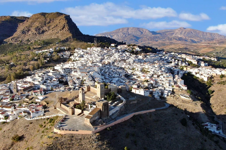 Aerial view of a hilltop fortress surrounded by a town with white buildings, set in a mountainous landscape under a blue sky.