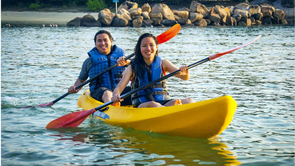 Two people in life vests paddle a yellow kayak on calm water, with rocks and trees on the shoreline in the background.