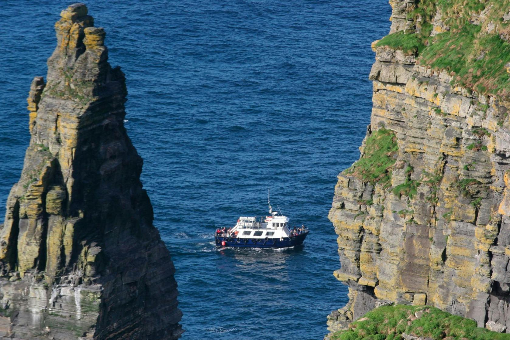 A white and blue boat sailing between two towering cliffs over a calm blue sea.