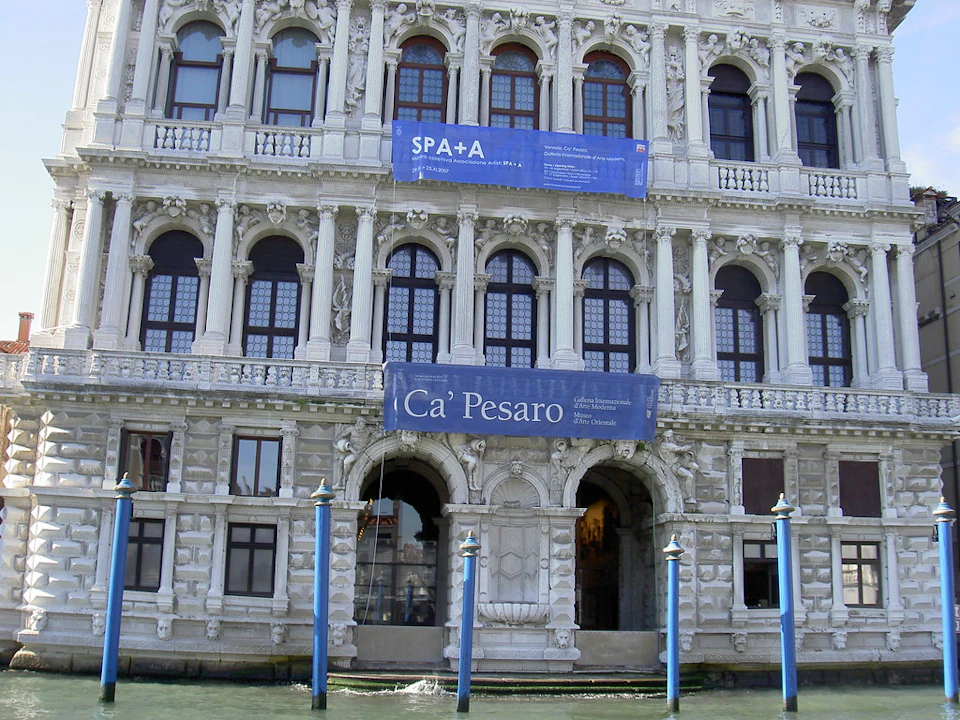 Ornate façade of Ca' Pesaro in Venice with two blue banners and blue poles in the canal below.