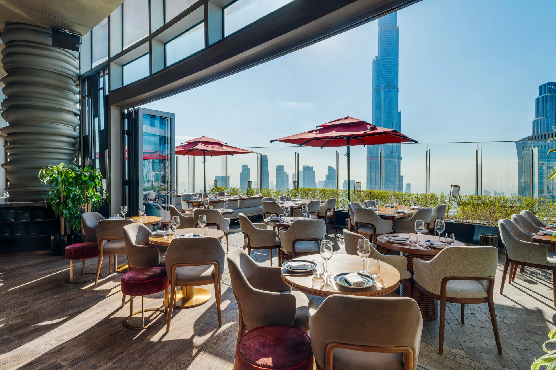 Modern outdoor restaurant with tables set for dining, red umbrellas, and a city skyline including a tall skyscraper in the background.