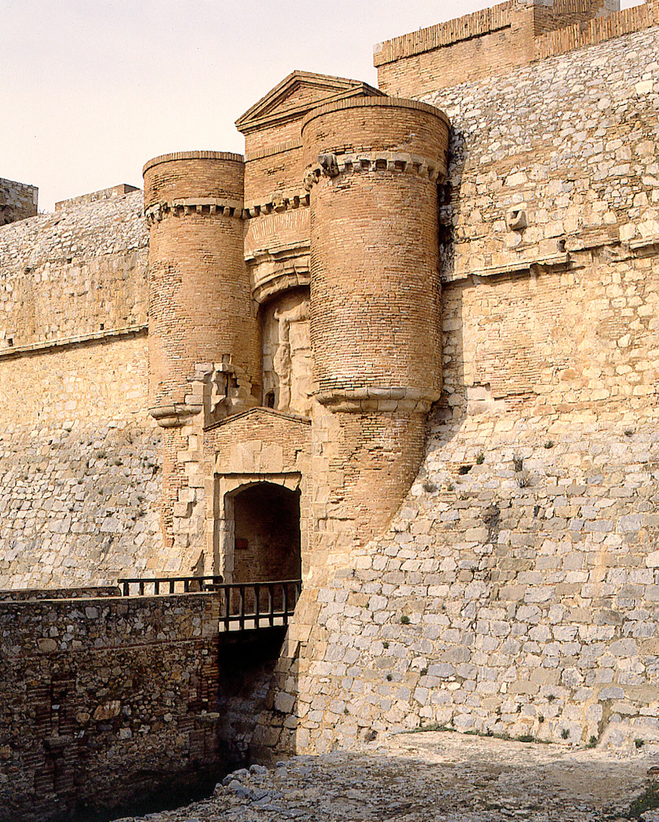Stone fortress with two large cylindrical towers and a gate. The structure is made of rough stones and bricks.