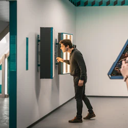 En person inspekterar en utställning på en vägg medan en annan fotograferar reflektioner i ett triangulärt spegelkalejdoskop.