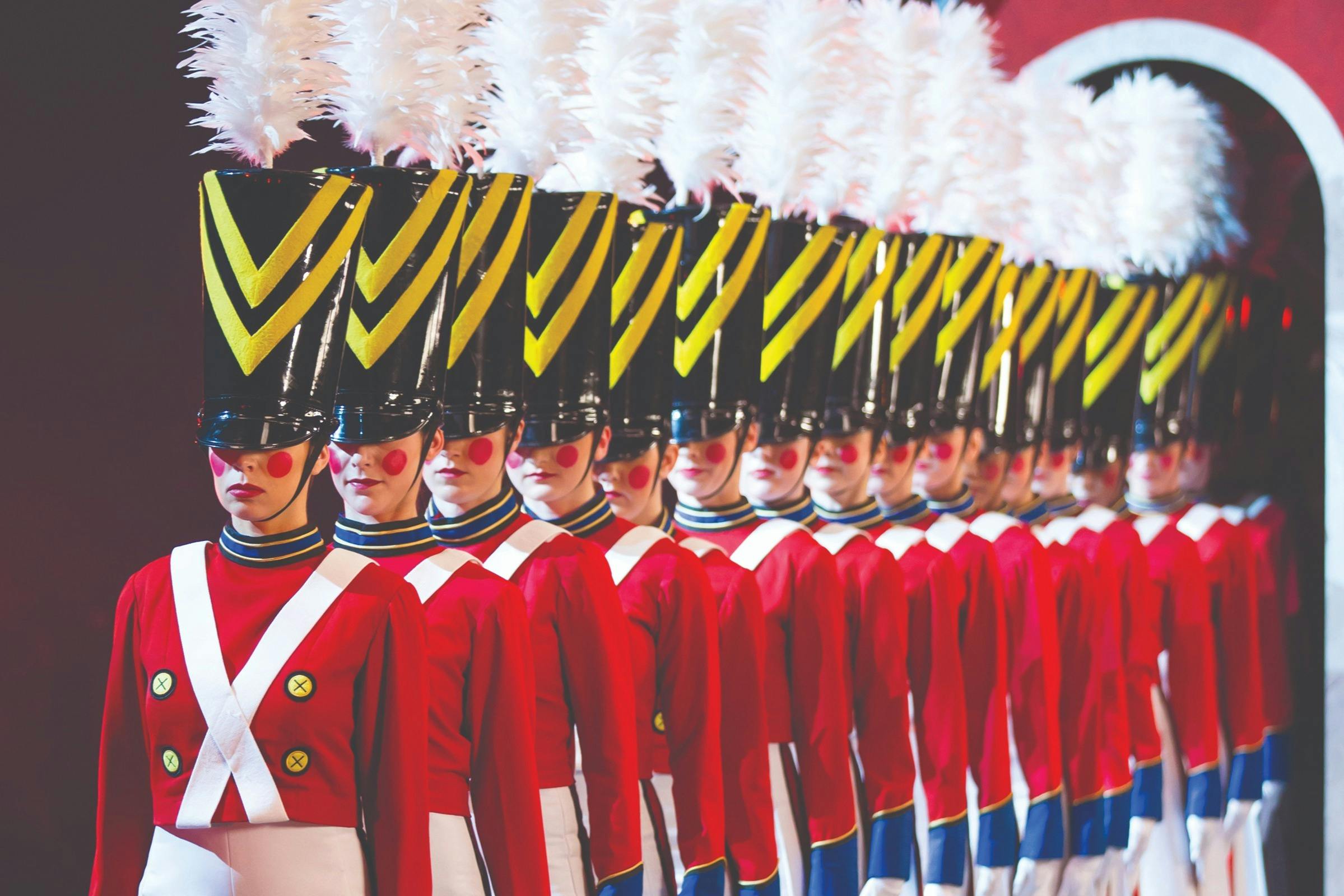 Řada účinkujících oblečených jako vojáčci s červenými uniformami, vysokými černými klobouky se žlutými prýmky a bílými chocholy.