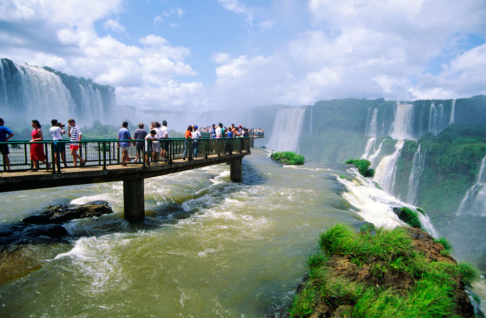 People stand on a viewing platform over a rapidly flowing river, with large waterfalls and lush greenery in the background.