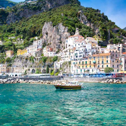 A small boat on turquoise water with colorful hillside buildings and lush greenery on the Amalfi Coast.
