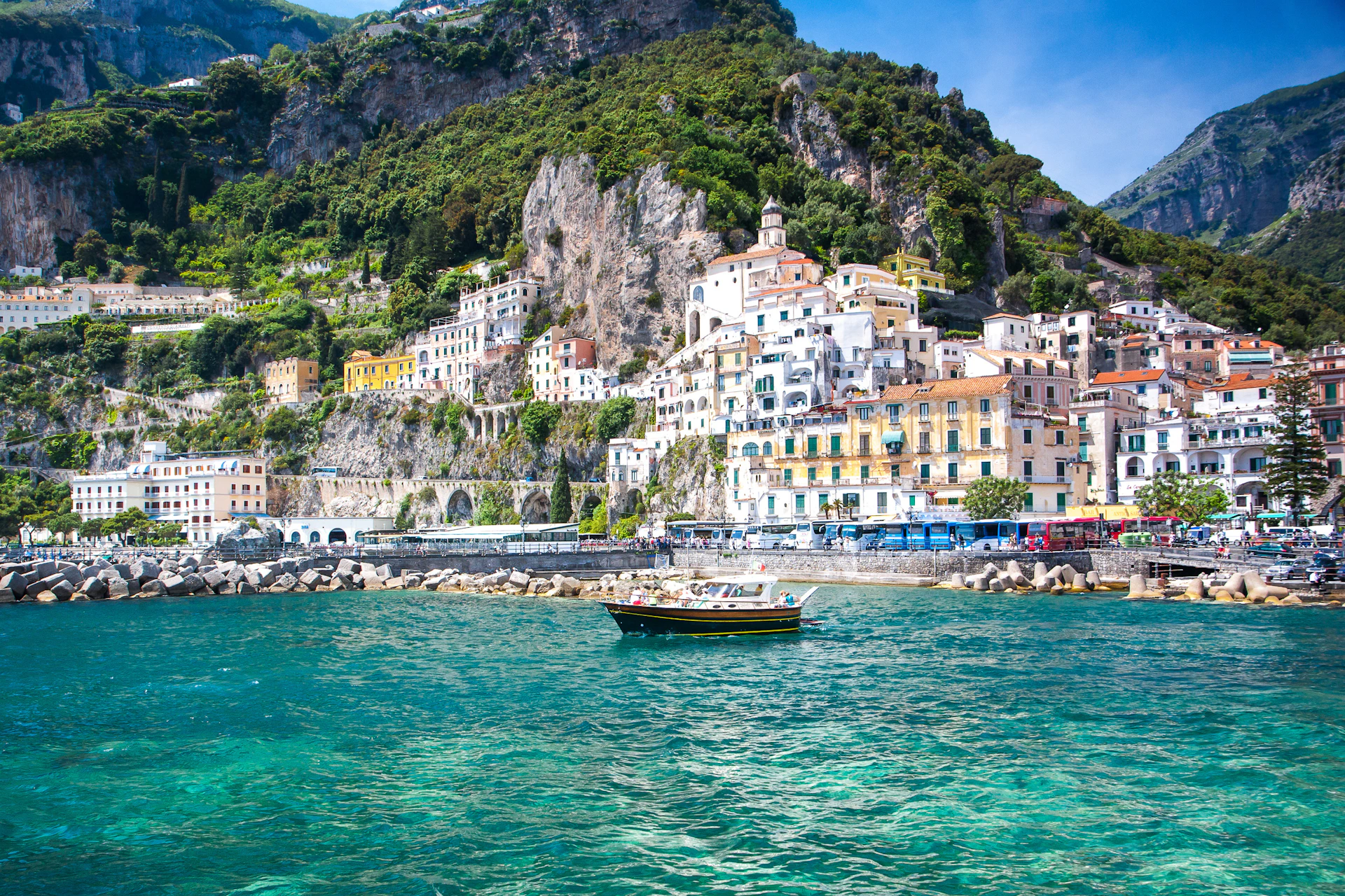 A small boat on turquoise water with colorful hillside buildings and lush greenery on the Amalfi Coast.