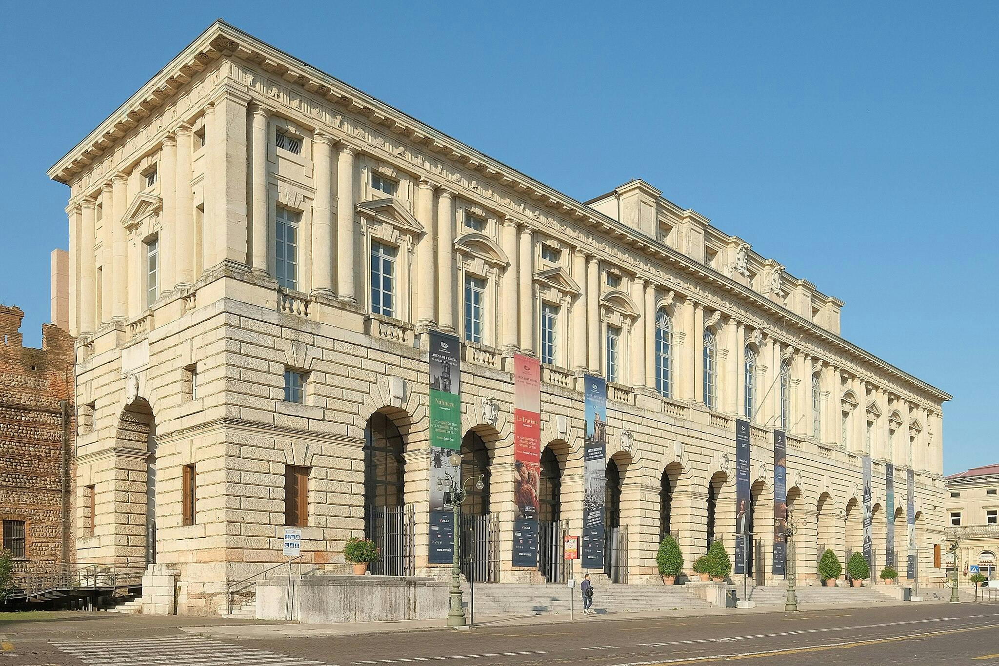 Bâtiment historique en pierre beige avec de hautes fenêtres cintrées, des bannières suspendues entre les colonnes et une personne marchant au premier plan.