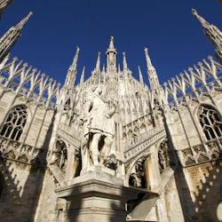 Gotisk katedralfasad med invecklade spiror och en staty av en klädd figur i mitten, under en klarblå himmel.