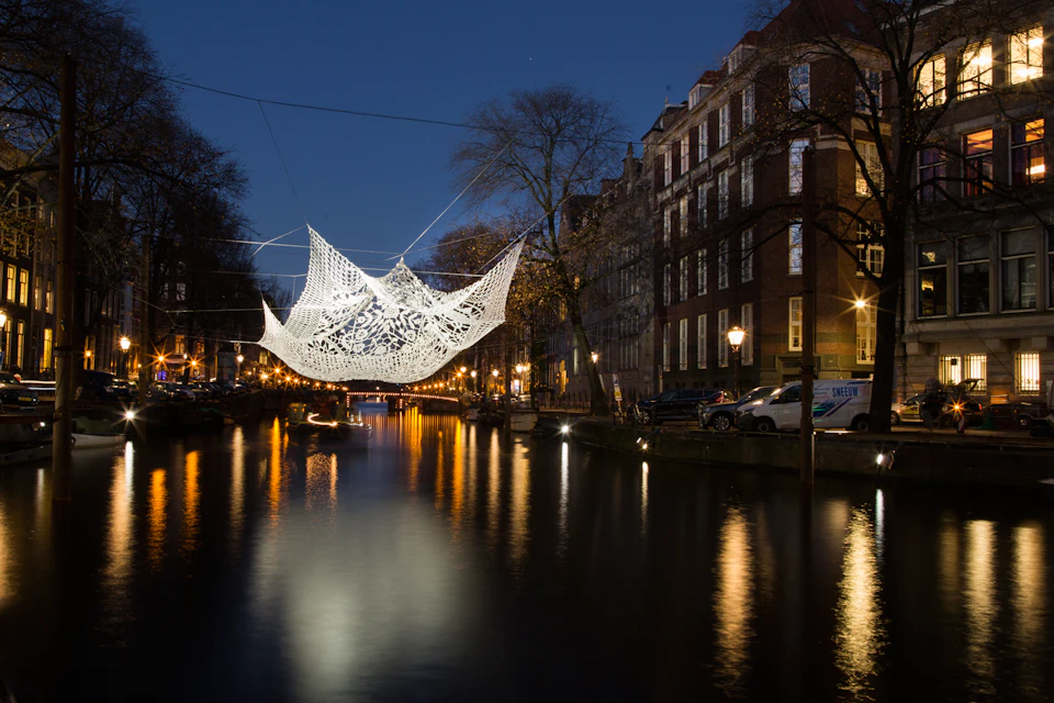 A luminous art installation hangs above a canal at night, surrounded by lit buildings and reflected lights in the water.