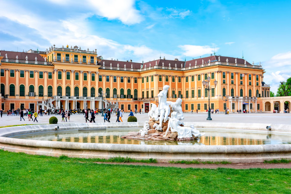 A grand yellow palace with multiple windows, an ornate fountain in the foreground, and people walking around the square.