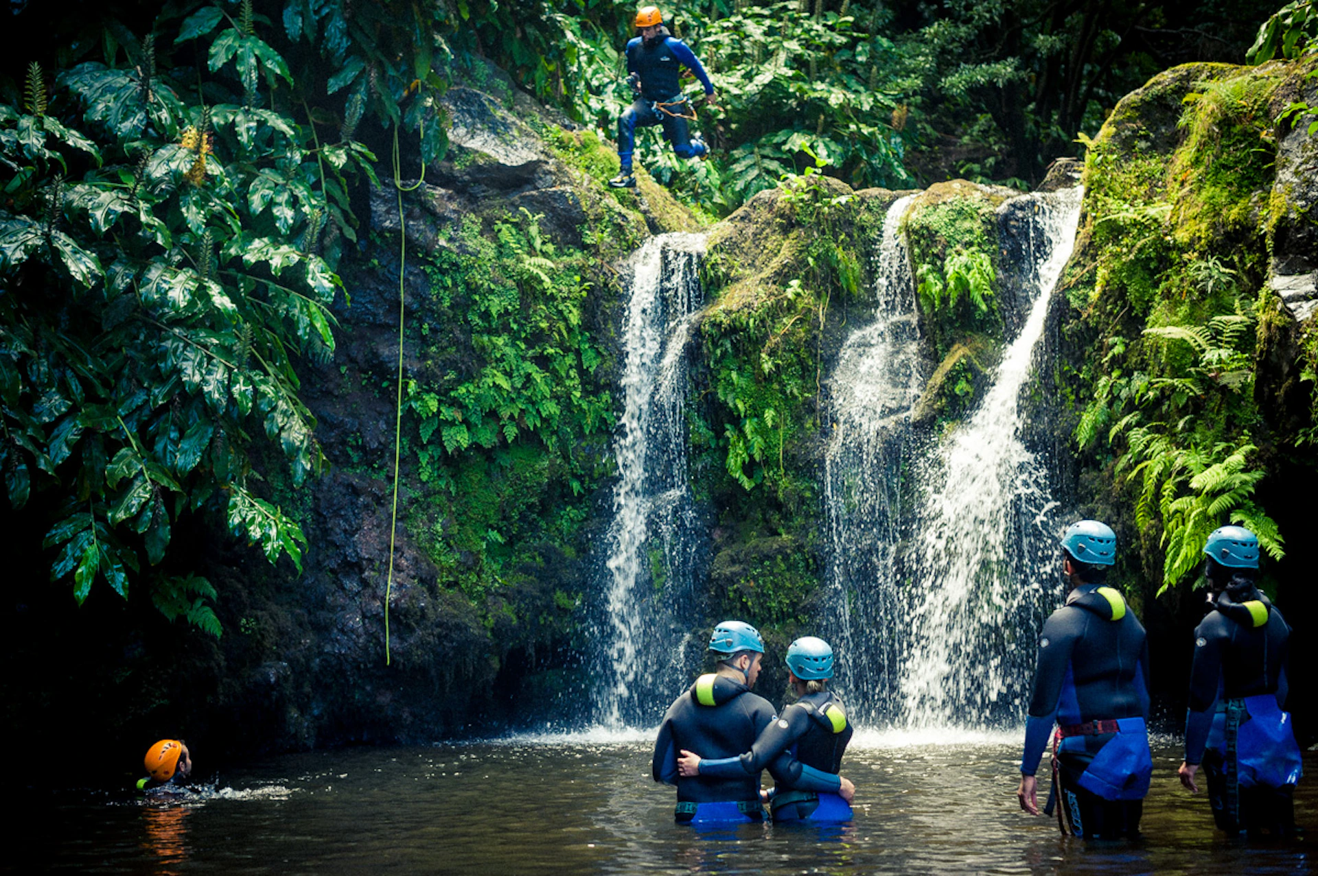 Two people in helmets stand in a pool below a mossy waterfall while another person jumps from the top, also wearing a helmet.